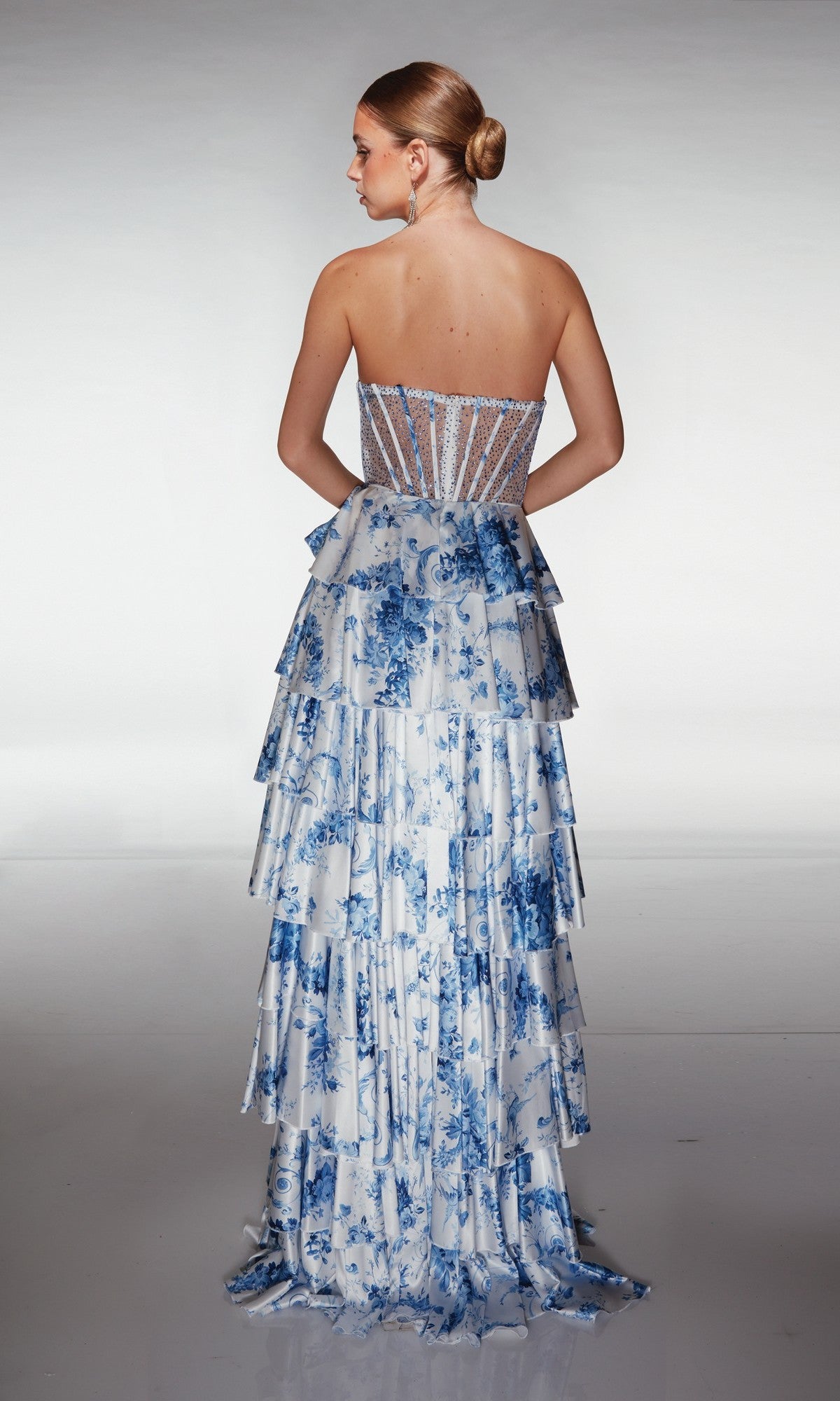 Woman wearing the Long Prom Dress: Aerie Couture 374, featuring blue and white florals, a sheer corset bodice, and a tiered ruffle A-line skirt, stands with her back to the camera against a plain backdrop.